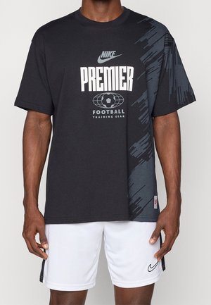 Man draagt een zwart Nike Premier voetbaltrainings-T-shirt met een afbeelding op de rechterschouder en witte Nike-shorts.
