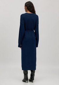 Navyblaues langärmliges Kleid mit entspannter Passform und tailliertem Gürtel, mit glatter Textur und knielangem Saum. Schwarze knielange Stiefel.