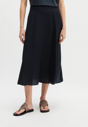 Femme portant une jupe midi noire et des sandales plates marron, debout devant un fond blanc uni.