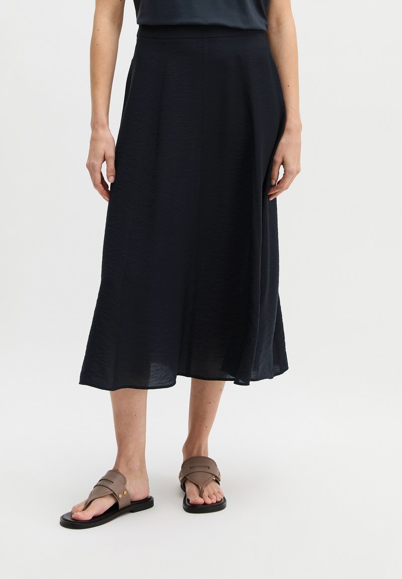 Vrouw draagt een zwarte midirok en bruine platte sandalen, staand tegen een effen witte achtergrond.