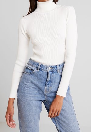 Vrouw draagt een wit geribd coltrui en lichtblauwe high-waisted denim jeans met voorzakken en riemlussen.
