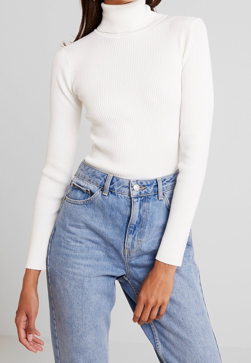 Femme portant un pull à col roulé côtelé blanc et un jean en denim bleu clair taille haute avec poches avant et passants de ceinture.