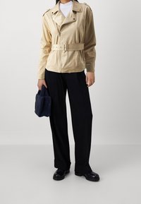 Personne portant une veste beige ceinturée, un pantalon noir à jambes larges, des bottes noires, tenant un petit sac à main texturé bleu marine, se tenant sur un fond uni.