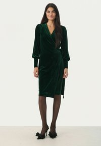 Grünes Samt-Wickelkleid mit langen Ärmeln, das eine gebundene Taillenbetonung und einen knielangen Saum hat. Getragen mit schwarzen, schuhen mit Schleifenakzenten.