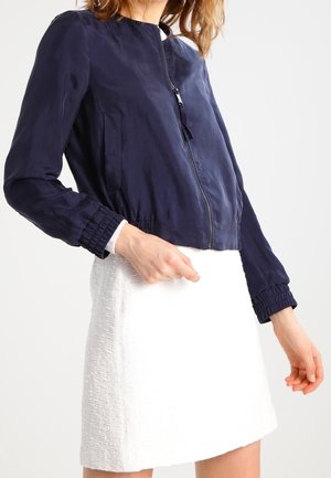 Kvinde iført en marineblå lynlåsjakke med opsamlede manchetter og en hvid tekstureret mininederdel, stående mod en ensfarvet baggrund.