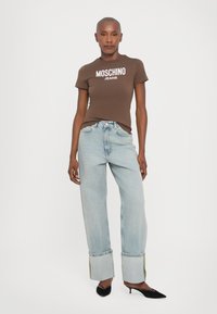 Brązowa dopasowana koszulka z logo "MOSCHINO JEANS", w połączeniu z jasnoniebieskimi spodniami wysokim w stan, podwiniętymi na kostkach, z złotymi akcentami. Czarne szpilki.