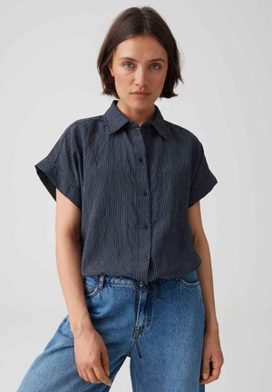 Donna con capelli corti castani che indossa una camicia blu scuro a maniche corte abbottonata e jeans azzurri su uno sfondo semplice.