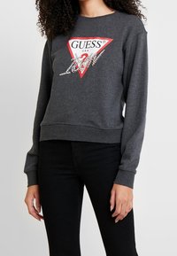 Mujer vistiendo un sudadera de Guess en gris oscuro con logo triangulo rojo y detalles de strass, combinada con pantalones negros, de pie frente a un fondo blanco.