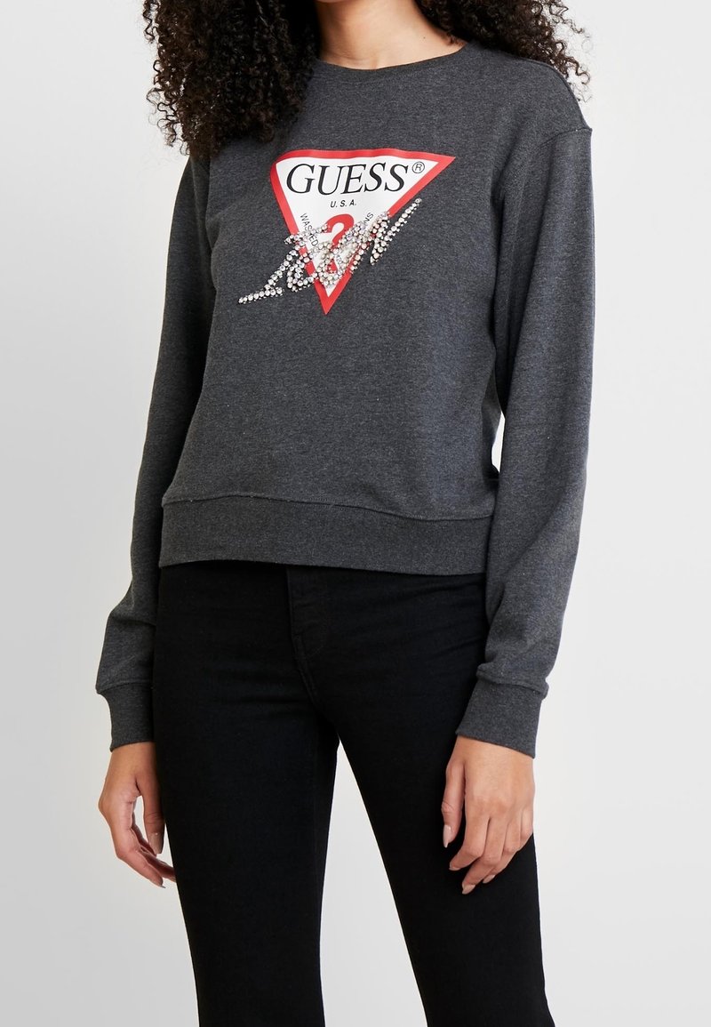 Mujer vistiendo un sudadera de Guess en gris oscuro con logo triangulo rojo y detalles de strass, combinada con pantalones negros, de pie frente a un fondo blanco.