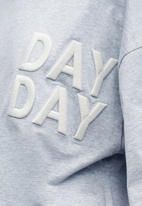 Ανοιχτό γκρι ύφασμα με ανάγλυφα λευκά κεντημένα γράμματα που σχηματίζουν τη φράση "DAY DAY" σε δύο σειρές.