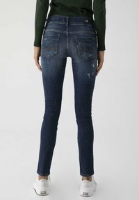 Jean skinny en denim bleu foncé avec des effets délavés clairs. Comprend deux poches arrière et une ceinture de marque. Porté avec des baskets blanches.