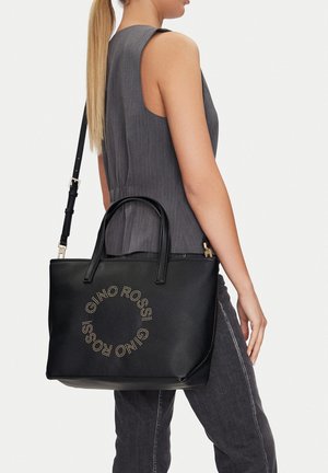 Mujer con coleta larga y rubia, vestida con top gris sin mangas y pantalones oscuros, llevando un bolso negro de Gino Rossi con un logo circular.