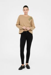 Maglione beige a maglia con dettagli a coste e decorazione floreale gialla sulla spalla, abbinato a jeans neri attillati e mocassini.