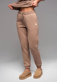 Beige, kurz geschnittene Sweatpants aus weichem Stoff mit elastischem Kordelzugbund und cuffs an den Knöcheln. Beigefarbene, rutschfeste Slipper mit Gummisohle.