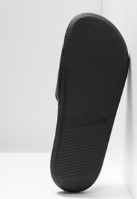 Semelle de sandale en caoutchouc noir avec des rainures diagonales et un logo de crocodile embossé, appuyée contre un mur blanc.