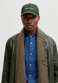 Green baseball cap with embroidered text, denim button-up shirt, houndstooth scarf with accents, and a dark leather jacket.