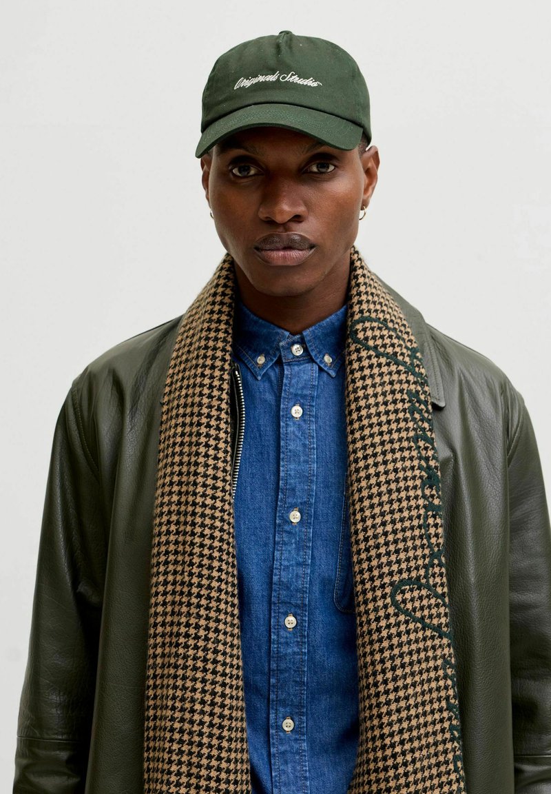 Green baseball cap with embroidered text, denim button-up shirt, houndstooth scarf with accents, and a dark leather jacket.