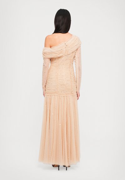 Vrouw met lang, donker haar die een perzikkleurige, off-shoulder, met pailletten versierde avondjurk draagt met een plooirok van tule, staande met de rug naar de kijker.