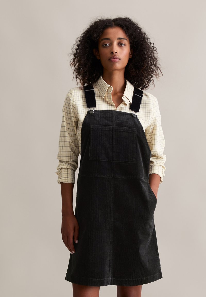 Robe salopette en velours côtelé noir avec bretelles réglables, poche avant et poches latérales, portée par-dessus une chemise à carreaux légère. Design simple.