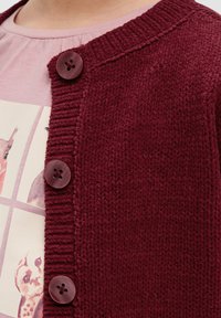 Bordeauxkleurige gebreide cardigan met een geribde kraag en vier ronde knopen, gedragen over een lichtroze graphic t-shirt met dierenprints.