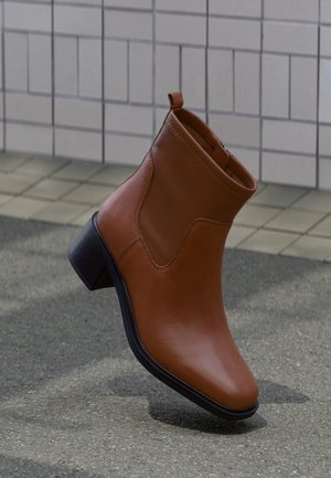 Brauner Lederknöchelstiefel mit niedrigem Blockabsatz auf grauem Pflaster und gefliester Wand im Hintergrund.