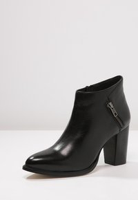 Zwarte leren enkelboot met spitse neus, blokhak en zijrits, tentoongesteld op een witte ondergrond.