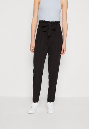 Kobieta nosząca czarne spodnie paperbag z wysokim stanem, zawiązanym paskiem i białe trampki, w zestawieniu z bezrękawnym jasnoszarym topem.