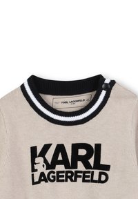 Beige gebreide trui met zwart-wit gestreepte ribbels in de kraag. Heeft een opvallend zwart "KARL LAGERFELD" logo. Knopdetail op de linkerschouder.