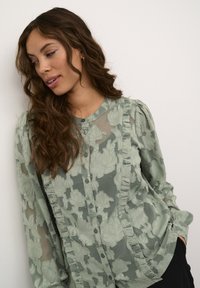 Blusa trasparente verde chiaro con ricami floreali, bordi arricciati e chiusura con bottoni. Maniche lunghe con dettagli di texture e motivo delicati.
