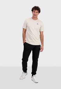 Beige bomull t-shirt med liten blå och röd logotyp på bröstet, tillsammans med svarta joggingsbyxor och vita sneakers. Enkel, avslappnad design.