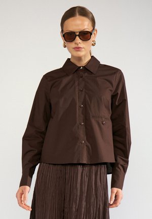 Woman wearing dark brown button-up shirt, matching pleated skirt, brown sunglasses, and gold hoop earrings, standing against plain background.