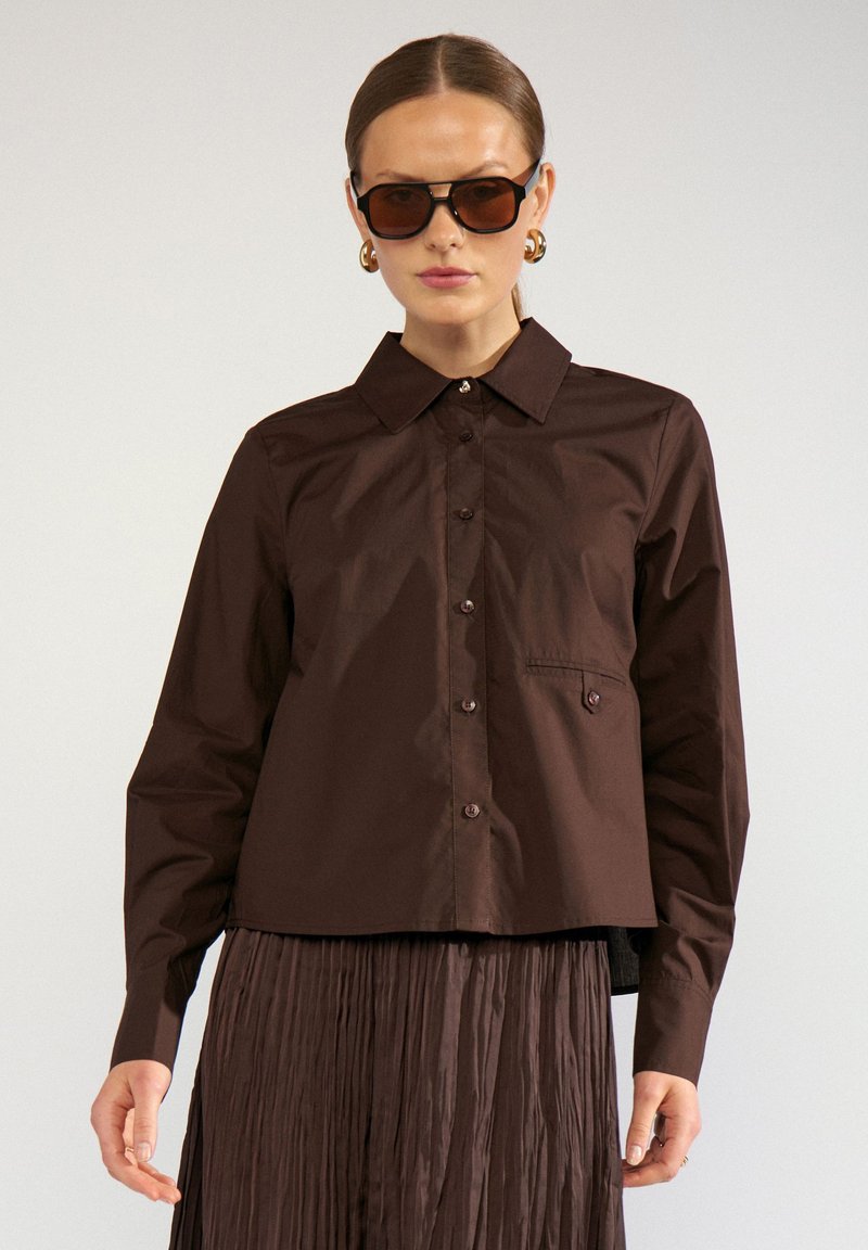 Femme portant une chemise marron foncé boutonnée, une jupe plissée assortie, des lunettes de soleil marron et des boucles d'oreilles créoles dorées, debout devant un fond uni.