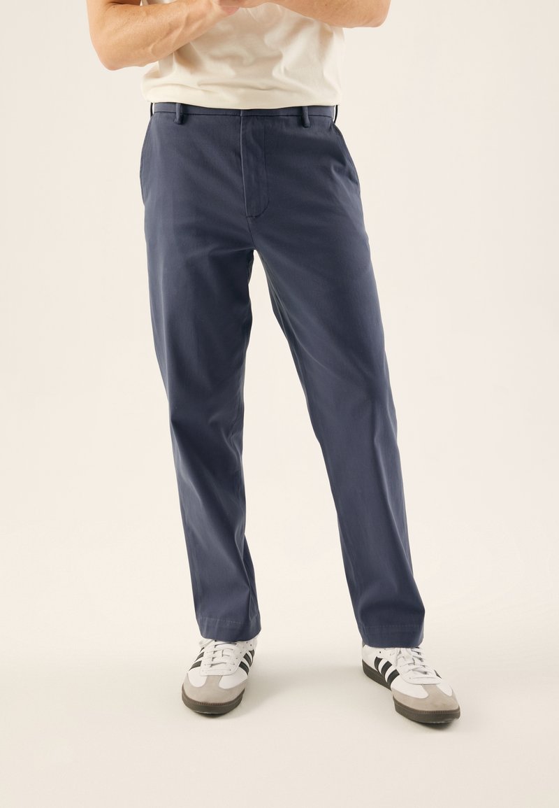 Donkerblauwe katoenen broek met rechte pijpen en een plooi aan de voorkant. Draagt witte en grijze sportschoenen met zwarte strepen.