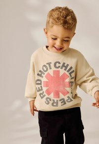 Uśmiechnięty młody chłopiec z kręconymi blond włosami, ubrany w beżowy sweter z czerwonym gwiazdką i napisem "Red Hot Chili Peppers", trzymający kogoś za rękę.