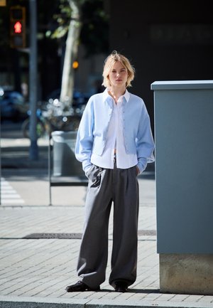 Lyseblå cardigan med knaplukning over en hvid skjorte, parret med løse grå bukser. Sorte sko synlige, urban indstilling.