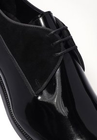 Chaussure en cuir noir avec une finition brillante, des accents en daim et des lacets ton sur ton. Le design comprend un bout arrondi et une silhouette structurée.