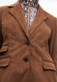 Hnědá semišová sako s klopovým límcem, dvěma předními kapsami a dvěma černými knoflíky. Vnitřní vrstva má texturovanou vzorovanou látku.