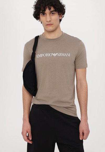 Beige cotton t-shirt featuring a white "Emporio Armani" logo, round neckline, short sleeves, and paired with black pants and a shoulder bag.