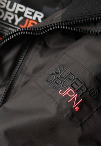 Czarna kurtka z wzorem w kratę, z zapięciem na zamek błyskawiczny oraz naszywką z logo "SUPER DRY JPN" w haftowanych czarnych i czerwonych literach.