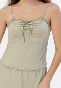 Camiseta de tirantes ligera en color verde claro con un diseño ajustado, bordes con volantes, tirantes ajustables y un detalle de lazo en el centro. Textura suave y lisa.