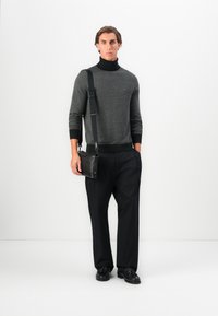 Hombre vestido con un suéter de cuello alto gris con patrones y pantalones negros, sosteniendo un bolso cruzado de cuero negro, de pie frente a un fondo blanco.