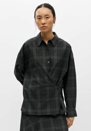 Jeune femme aux cheveux foncés attachés porte une chemise à carreaux gris foncé boutonnée et une jupe assortie sur fond blanc.