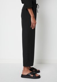 Zwarte, loose-fit broeken met een elastiche tailleband en zijzakken, gecombineerd met zwarte sandalen met kruisbanden, van opzij gefotografeerd.
