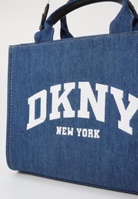 Džinsinė tote rankinė su dviem trumpais rankenomis, kurią puošia ryškūs baltos spalvos "DKNY" užrašai ir mažesnis "NEW YORK" šriftas. Matomas kontrastingas siuvinėjimas.