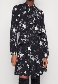 Musta mekko, jossa on valkoinen tähtikuosi, rypytetty kaula-aukko, pitkät hihat nappikiinnityksellä ja vyönauha vyötäröllä, teksturoitu kangas.