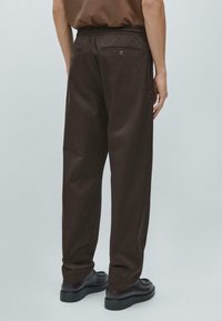 Brązowe, dopasowane spodnie z elastyczną talią, prostymi nogawkami i dwoma tylnymi kieszeniami. Gładka tekstura z delikatnym połyskiem, zestawione z czarnymi butami.