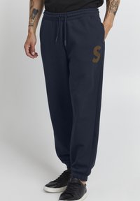 Marineblå joggebukser med elastisk midjebånd, sidelommer og en brun "S" logo. Laget av mykt stoff med innsnevrede mansjetter. Svarte sneakers.