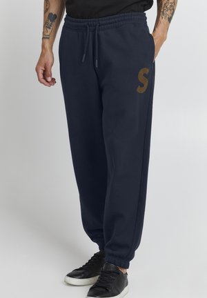 Navy sweatpants with an elastic waistband, side pockets, and a brown "S" logo. Made of soft fabric with tapered cuffs. Black sneakers.
