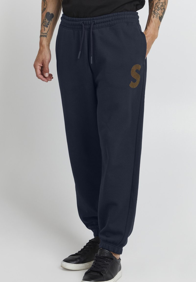 Marineblå joggebukser med elastisk midjebånd, sidelommer og en brun "S" logo. Laget av mykt stoff med innsnevrede mansjetter. Svarte sneakers.