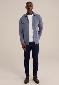 Lichtblauwe button-up shirt met twee borstzakken, gedragen over een wit T-shirt, gecombineerd met donkerblauwe jeans en bruine veterschoenen.